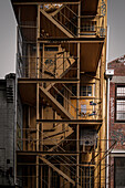 View of a yellow metal fire escape clinging to the side of an old building, a stark contrast against the muted brick and grey skies, Hobart, Tasmania, Australia.