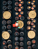 Luftaufnahme einer Person mit rotem Kegelhut inmitten von Reihen von Steinguttöpfen, die ein Muster aus rustikalen Braun- und Erdtönen bilden, Sojasoßenhersteller, Dorf Ban Yen Nhan, Hanoi Vietnam.