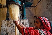 Bogura, Bangladesh - 24 April 2021: View of a woman in a red patterned sari, carefully handling strands of noodles emerging from an old, blue and beige machine in a factory.