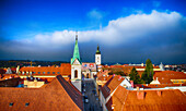 View of an old town square with red-tiled roofs and a green-spired Church of St. Mark under a vibrant blue sky with fluffy white clouds, Zagreb, Croatia.