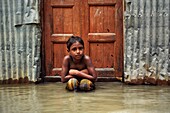 Bogura, Bangladesh - 15 July 2019: View of a young boy, his eyes reflecting the somber mood, sits amidst floodwaters against corrugated iron and a weathered wooden door.