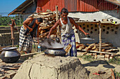Feni, Bangladesh - 26 November 2018: View of two men cooking outdoors on a rustic stove, steam rising against the backdrop of corrugated iron and stacked wood.