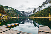 Blick auf den ruhigen See, in dem sich majestätische, in Nebel gehüllte Berge spiegeln, eingerahmt von hölzernen Stegen, die eine ruhige Szene schaffen, Gousau, Österreich.