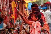 Bogura, Bangladesh - 03 November 2023: View of a young girl's focused gaze amidst the vibrant, colorful necklaces at a bustling market stall, contrasting with the muted tones of the background crowd.