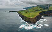 Luftaufnahme der dramatischen Klippen, die auf den weiten Ozean treffen und den Kontrast zwischen den dunklen Felsen und den üppigen grünen Feldern darüber zeigen, Santa Cruz das Flores, Portugal.