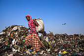 Chittagong, Bangladesch - 08. März 2024: Blick auf eine Frau in einem rot-weiß gemusterten Kleidungsstück, die auf eine riesige Mülldeponie klettert und einen schweren Sack gegen einen klaren blauen Himmel trägt.