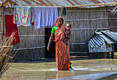 Bogura, Bangladesch - 07. März 2020: Blick auf eine Mutter, die durch die Fluten watet und ihr Kind neben einer bescheidenen Behausung aus Stroh und Blech in den Armen hält - ein beeindruckendes Bild der Resilienz.