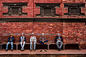 Dhangadhi, Nepal - 27 February 2023: View of men in traditional Nepali attire sit in a row against the backdrop of an old brick building with ornate windows.