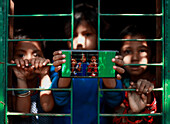 Mirsharai, Bangladesch - 16. Januar 2019: Blick auf Kinder, die durch ein grünes, vergittertes Fenster spähen, eines hält ein Telefon, auf dem sie sich spiegeln.