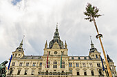 View of city hall, ornate building with flags, a tower, and a tall decorated pole reaching towards a cloudy sky, Graz, Austria.