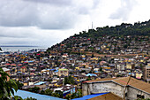 Freetown, Sierra Leone - 11. August 2021: Blick auf eine ausgedehnte Stadtlandschaft, die sich über einen üppigen Hügel zum schimmernden Ozean hinunterzieht, ein buntes Geflecht von Dächern unter einem bewölkten Himmel.