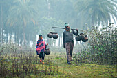 Natore, Bangladesch - 17. Januar 2023: Blick auf einen Mann und ein Kind, die inmitten der gedämpften Töne des Winters die nebligen Felder durchqueren und irdene Töpfe auf einem Joch tragen.