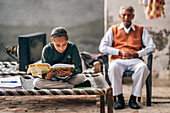 Kolkata, India - 28 January 2023: View of a young girl engrossed in studies on a traditional cot, with a senior man watching over her in the warm daylight.