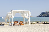 View of sun-drenched beach cabana with billowing white curtains frames a serene turquoise sea, inviting relaxation and escape, Lindos, Rhodes, Greece.