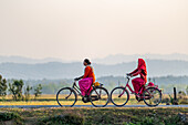 Dhangadhi, Nepal - 03 March 2023: View of two women in vibrant red and orange traditional clothing cycling along a road, framed by a backdrop of distant hills.