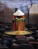 Hanoi, Vietnam - 09. April 2024: Ansicht eines Arbeiters mit traditionellem Kegelhut, der goldene Körner über Reihen von Tonkrügen siebt, die eine Szene von rustikaler Industrie darstellen.