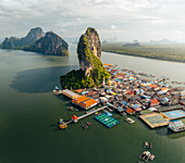 Aerial view of stilt houses clinging to the base of a towering karst formation in Phang Nga Bay, a spectacle of human ingenuity and natural wonder, Ko Panyi, Phang Nga, Thailand.