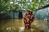 Bogura, Bangladesch - 15. Juli 2019: Blick auf eine Mutter und ein Kind, die in der Nähe von Wellblechkonstruktionen durch die Fluten waten, in denen sich der bedeckte Himmel und das dichte Laubwerk in einer düsteren Szene spiegeln.