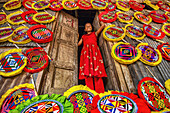 Gaibandha, Bangladesh - 04 September 2020: View of a girl in red attire smiles from a doorway adorned with colorful, handcrafted fans, creating a vibrant display of local artistry.