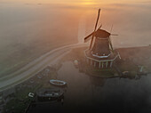 Aerial view of a windmill shrouded in a mystical fog, the golden sun casting a warm glow over the tranquil waters, Schansend, Noord-Holland, Netherlands.
