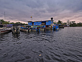 View of vibrant blue buildings and boats reflecting in the dark, rippled water under a cloudy sky, creating a tranquil river scene, Guainia, Guainia, Colombia.