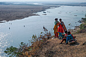 Sunamganj, Bangladesh - 27 January 2019: View of women in vibrant attire overlooking the shimmering Surma River, its waters reflecting the pale sky and distant sandbanks.