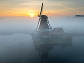 Aerial view of a majestic windmill shrouded in mystical fog, reflecting in the tranquil waters at sunrise, Schansend, Noord-Holland, Netherlands.