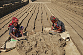 Feni, Bangladesh - 26 November 2018: View of two young girls diligently working with mud, shaping bricks under the harsh sun, their vibrant headscarves contrasting with the monotonous landscape.