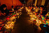 Dhaka, Bangladesh - 13 November 2018: View of rows of people seated beside a wall, illuminating the night with glowing diyas, their warm light reflecting on faces and the dark pavement.