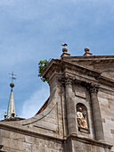 Blick auf ein altes, steinernes Gebäude mit einer Statue in einer Nische unter einem teilweise bewölkten Himmel, ergänzt durch einen Kirchturm mit einem Kreuz, Alcala de Henares, Gemeinde Madrid, Spanien.