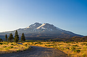 Blick auf den majestätischen, schneebedeckten Mount Shasta, der sich über ein goldenes Feld erhebt. Eine unbefestigte Straße schlängelt sich durch die sonnenüberflutete Landschaft, Mount Shasta, Kalifornien, Vereinigte Staaten.