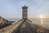 View of a stone walkway leading to the Phare de Kermorvan lighthouse under a serene sky as the sun sets over the ocean, Le Conquet, Bretagne, France.
