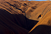 Blick auf den kolossalen, sonnenverbrannten Uluru-Monolithen, der tiefe Schatten wirft und die strukturierte, orange-rote Felswand offenbart, Lasseter Highway, Uluru, Northern Territory, Australien.