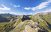 Blick auf zerklüftete Berggipfel, die sich unter einem strahlend blauen Himmel mit weißen Schäfchenwolken erheben und deren Hänge in Grün und Braun gefärbt sind, Schareck, Tirol, Österreich.