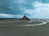 Luftaufnahme des Mont Saint-Michel, der sich unter einem düsteren Himmel majestätisch aus dem Wattenmeer erhebt, seine alten Steine sind ein Zeugnis der Zeit, Mont Saint-Michel, Normandie, Frankreich.