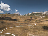 Luftaufnahme einer kurvenreichen Straße, die sich durch die goldene, sonnenüberflutete Hochebene schlängelt und zu fernen, schneebedeckten Bergen unter einem weiten, azurblauen Himmel führt, Gran Sasso, Abruzzen, Italien.