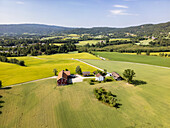Aerial view of a rustic red farmhouse nestled amidst vibrant green and yellow fields, contrasting against the backdrop of forested hills, Heddal, Telemark, Norway.