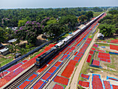 Aerial view of a vibrant red chili harvest drying alongside a train track under the sun, creating a striking contrast between rural life and modern transportation, Bogura, Bangladesh.