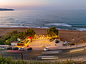 Aerial view of warm lights spill across the beachside bar as dusk settles over the undulating waves, cars blur along the coastal road, Chania, Chania, Greece.