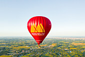 Sint-Martens-Latem, Belgium - 25 August 2025: Aerial view of a vibrant red hot air balloon soaring above the lush green landscape, a beacon against the soft, blue sky.