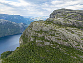 Aerial view of the dramatic Preikestolen cliff face rising above the deep blue Lysefjord waters, its rugged texture contrasting with the distant misty mountains, Lysefjord, Vestland, Norway.