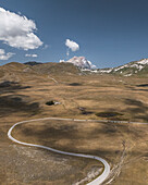 Luftaufnahme der kurvenreichen Straße, die eine blasse Schneise durch das goldene Grasland schlägt und zu den fernen, schneebedeckten Gipfeln des Gran Sasso führt, Campo Imperatore, Abruzzen, Italien.