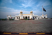 View of a white building with gold domes under a bright sky next to a waving flag, with colorful vehicles parked nearby, Kartarpur, Punjab, Pakistan.
