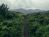 Aerial view of railroad tracks cutting through verdant jungle towards distant mountain peaks under a cloudy sky, Neturlim, Goa, India.