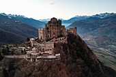 Luftaufnahme von Sacra di San Michele, einem mittelalterlichen architektonischen Wunderwerk auf einem Berg, dessen antike Steine in das sanfte Licht der Dämmerung getaucht sind, Sacra di San Michele, Piemonte, Italien.