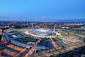 Turin, Italy - 21 August 2022: Aerial view of the Juventus Stadium, a modern architectural marvel, glows warmly against the fading twilight, a beacon amidst the sprawling urban landscape.