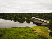 Aerial view of a bridge standing tall over the tranquil Nemunas River, reflecting the gray sky above against the backdrop of deep green forests, Merkine, Lithuania.