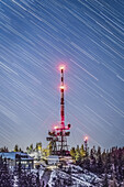 Blick auf einen rot-weißen Fernmeldeturm auf dem Schöckl, der den Nachthimmel durchdringt, während die Sterne über ihm die Landschaft am Schöckl in der Steiermark, Österreich, erhellen.