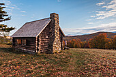 Blick auf eine rustikale Hütte mit steinernem Schornstein auf einem Hügel inmitten des bunten Herbstlaubs unter einem strahlenden Himmel, mit einem kleinen Kind, das auf das Feld hinausschaut. White Mountains, New Hampshire, Vereinigte Staaten.