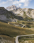 Luftaufnahme der kurvenreichen Straße, die sich durch die goldenen Felder in Richtung der majestätischen Berggipfel schlängelt, Gran Sasso, Abruzzen, Italien.